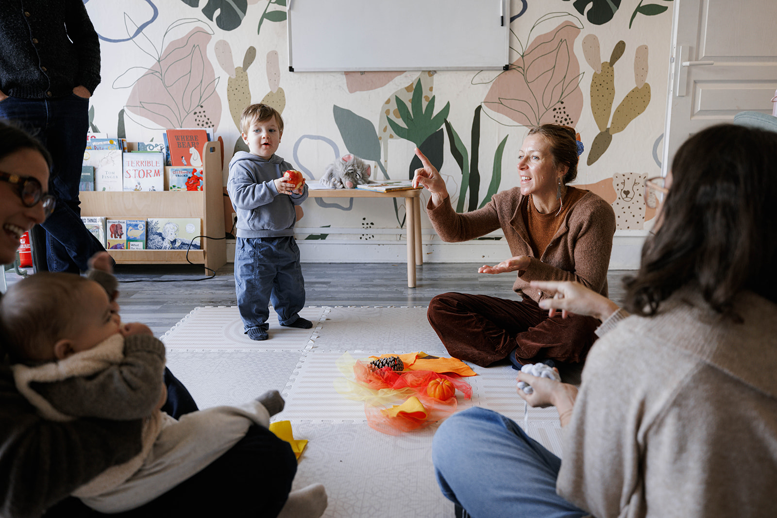 Two fair-skinned, young adults converse with two babies and a young boy, while sitting on a white mat in a room filled wit...