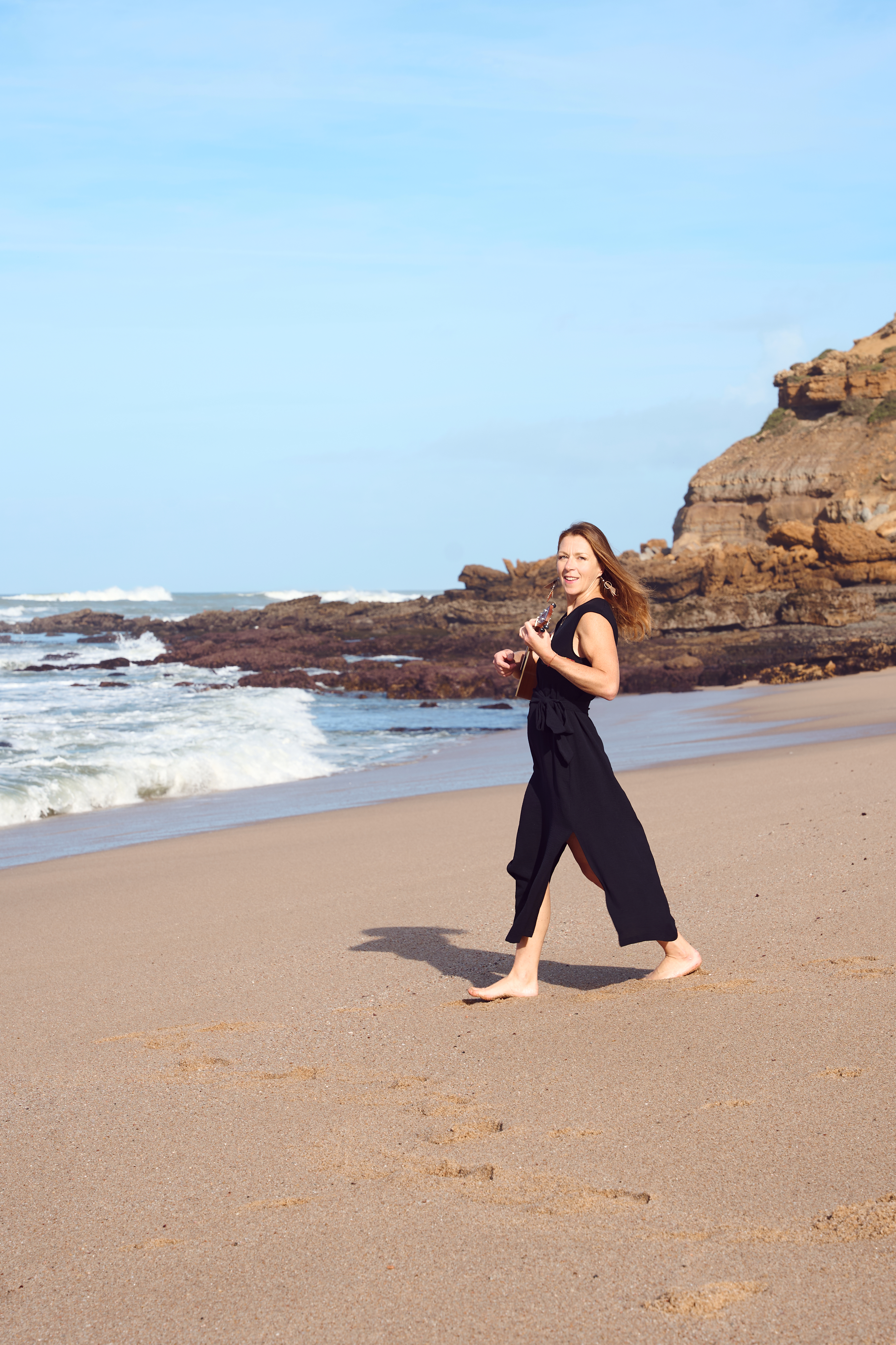 The image depicts a young woman in black attire strolling along a sandy beach, with the ocean waves gently lapping at her ...